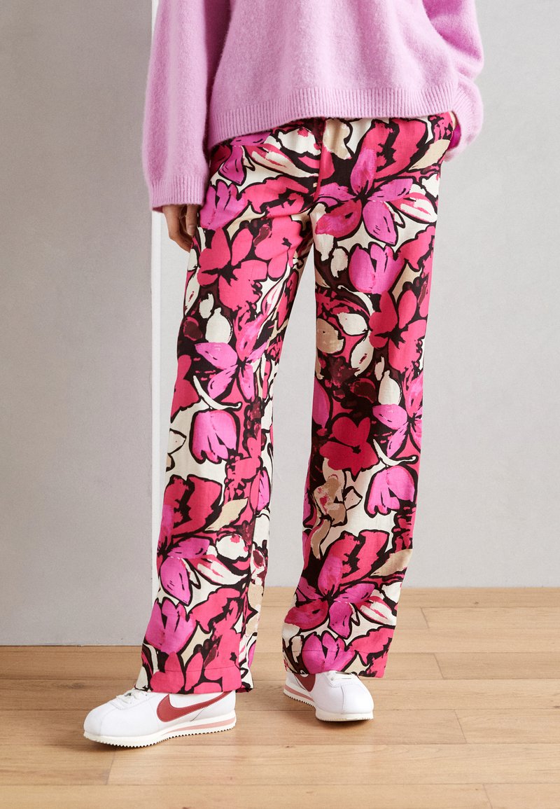 Blommönstrade vida byxor i livliga nyanser av pink, röd och beige, kombinerade med vita sneakers och en rosa tröja. Slät tygtextur.