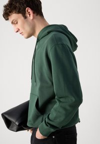 Felpa verde in cotone con tasca frontale, coulisse e polsini a costine. Indossata con pantaloni neri e una borsa nera. Design semplice e pulito.