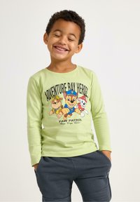 Lichtgroen T-shirt met lange mouwen en een grafische afbeelding van drie cartoonhonden. Gecombineerd met donkergrijze broek met zakken en een gestructureerd oppervlak.