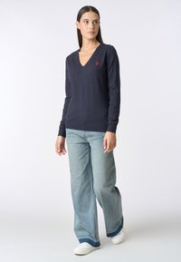 Maglione a V blu navy in morbido tessuto a maglia, con maniche lunghe e un piccolo logo rosso. Abbinato a jeans a gamba larga blu chiaro e sneakers bianche.