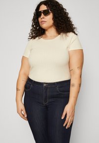 Top cream com texturização e mangas curtas, combinado com jeans azul-escuro. Os jeans possuem fecho por botão e um ligeiro desgaste, com arte corporal visível no braço.