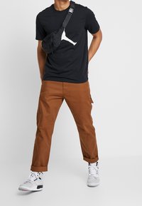 Camiseta negra con gráfico blanco; pantalones cargo marrones; bolso cruzado negro; zapatillas grises y blancas con detalles; puños doblados en los pantalones.