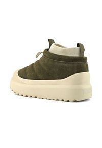 Zapato deslizante de ante verde oliva con suela gruesa beige, puño de punto acanalado en el tobillo y lengüeta en el talón, en un ángulo que muestra el lado exterior y la parte trasera.