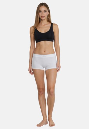 Woman with long hair wearing a black sports bra and white cotton sport boyshort underwear, standing barefoot against a white background.