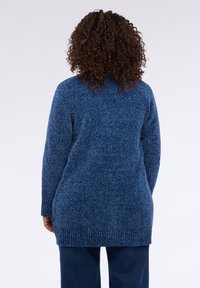 Persona con cabello rizado viste un suéter largo de punto azul y pantalones azul oscuro, de pie de espaldas frente a un fondo liso.
