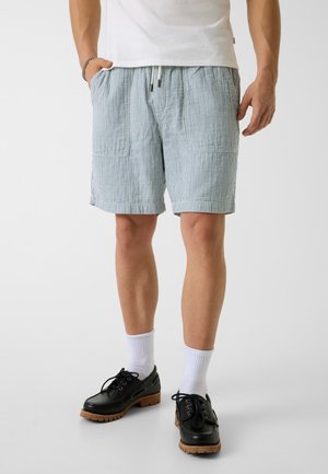 Mężczyzna ubrany w jasnoniebieskie i białe szorty w paski z troczkami, białe skarpetki sięgające nad kostkę, czarne skórzane sznurowane buty i białą koszulkę.