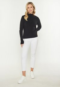 Schwarzer gerippter Rollkragenpullover, eng anliegend, kombiniert mit weißen engen Jeans und weißen Sneakers. Vor einem hellen Hintergrund, der eine Ganzkörperansicht zeigt.