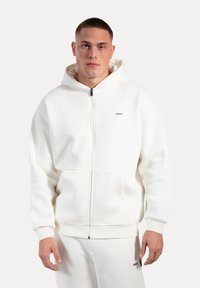 Hvid zip-up hoodie lavet af blødt stof, med en kængurulomme, ribbede manchetter og et lille logo på brystet. Glat tekstur.