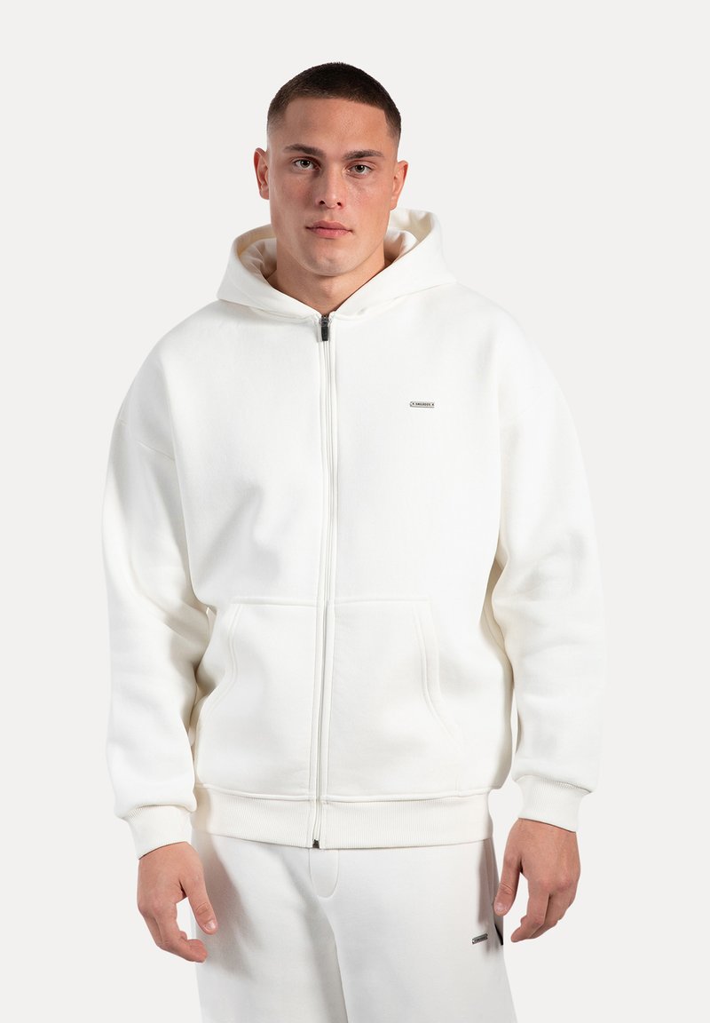 Sudadera blanca con cremallera hecha de una tela suave, que cuenta con un bolsillo tipo canguro, puños de canalé y un pequeño logo en el pecho. Textura suave.
