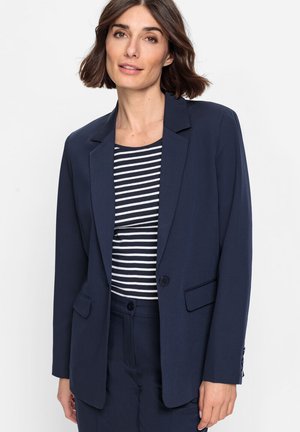 Femme aux cheveux courts et bruns portant un blazer bleu marine sur une chemise rayée noir et blanc et un pantalon bleu marine assorti, sur un fond uni.