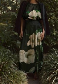 Maxikleid mit schwarzem Grund, das große weiße Blumen und grüne Blätter zeigt; elastischer Kurzschnitt in der Taille; kombiniert mit einem dunklen Blazer.
