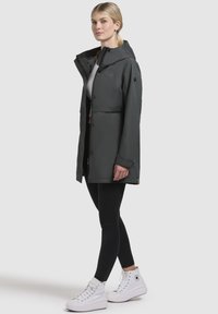 Donkergrijze waterdichte jas met capuchon, voorzien van een rits aan de voorkant en drukknoopsluiting, gedragen over zwarte leggings en witte hoge sneakers.
