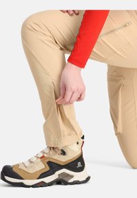 Beige cargobroek met ritsen en zijzakken, gecombineerd met tan en zwarte wandelschoenen met veters en een rubberen zool.