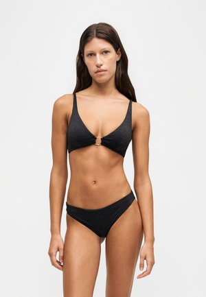Jonge vrouw met lang donker haar, gekleed in een zwarte bikini met een metalen ringdetail op het topje, staand tegen een eenvoudige lichte achtergrond.