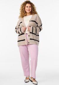 Bež pletena jopica z črnimi črtami, sprednjimi gumbi in dolgimi rokavi; v kombinaciji s svetlo roza hlačami in črno-belimi čevlji.