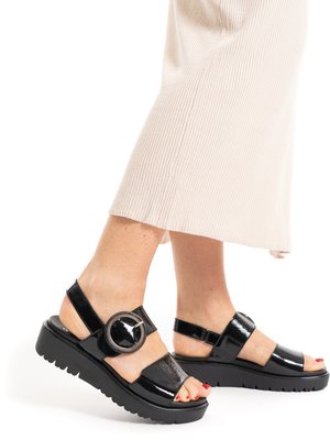 Feet wearing black patent leather platform sandals with large round buckle, paired with cream ribbed midi skirt.
