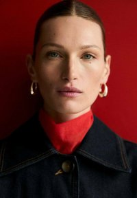 Portrait d'une personne portant une veste en denim foncé avec un col montant rouge. Elle a des boucles d'oreilles en argent en forme de hoop et un fond neutre.
