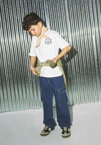 Camiseta blanca con gráfico de "COMIDA RÁPIDA", bandana verde, jeans cargo azules con bolsillos laterales, zapatillas marrones y negras, en interiores con fondo metálico.