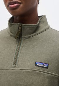 Κοντινό πλάνο ενός ατόμου που φοράει ένα πράσινο μπουφάν fleece Patagonia με ημι-φερμουάρ και λογότυπο στο στήθος.