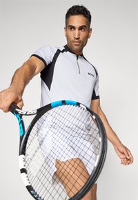 Mann in weißer und schwarzer Sportkleidung, der einen Tennisschläger nach vorne hält und sich vorbereitet, einen Schlag gegen einen einfarbigen Hintergrund zu machen.