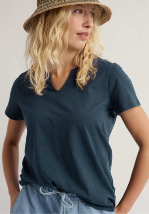 Mujer rubia con sombrero de paja, camiseta azul marino de cuello en V y pantalones azul claro con cordón, mirando de lado con las manos en las caderas.