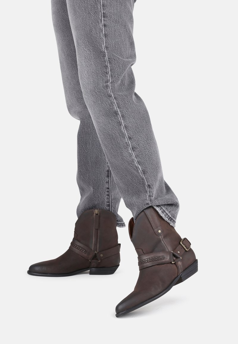 Botas de cuero marrón hasta el tobillo con punta afilada, cremallera lateral y hebillas decorativas. Combinadas con jeans de mezclilla grises, con un acabado texturizado.