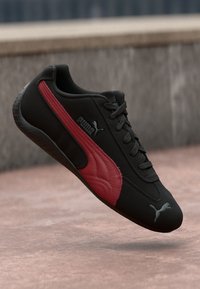 Fekete sportcipő textúrált piros oldalsó panellel, fűzős kialakítással és párnázott talppal. Puma logó az oldalon és az orrán.