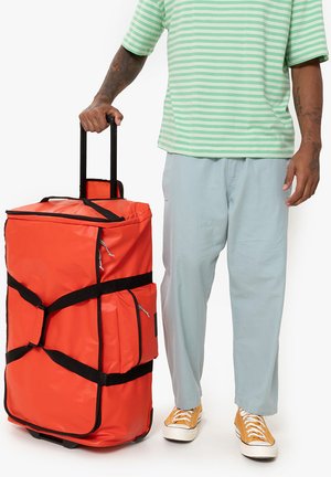 Red wheeled duffel bag with black straps and zippers, featuring a side pocket and a rectangular shape, held by a person in light pants.