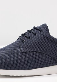 Marinblå skor gjorda av vävt tyg, med en texturerad yta, runda snören och en vit gummisula med en lätt kurva.