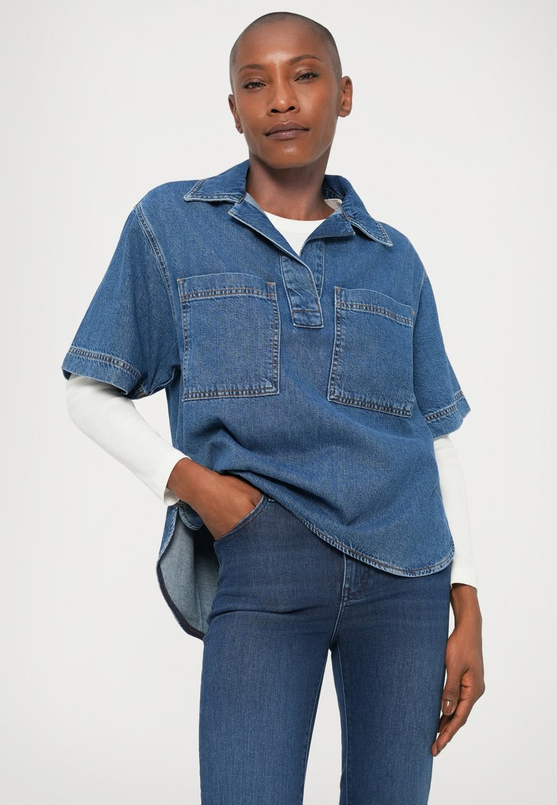 Denim shirt in blue with short sleeves, collar, two front pockets, and a side split hem. Worn over a white long-sleeve shirt.