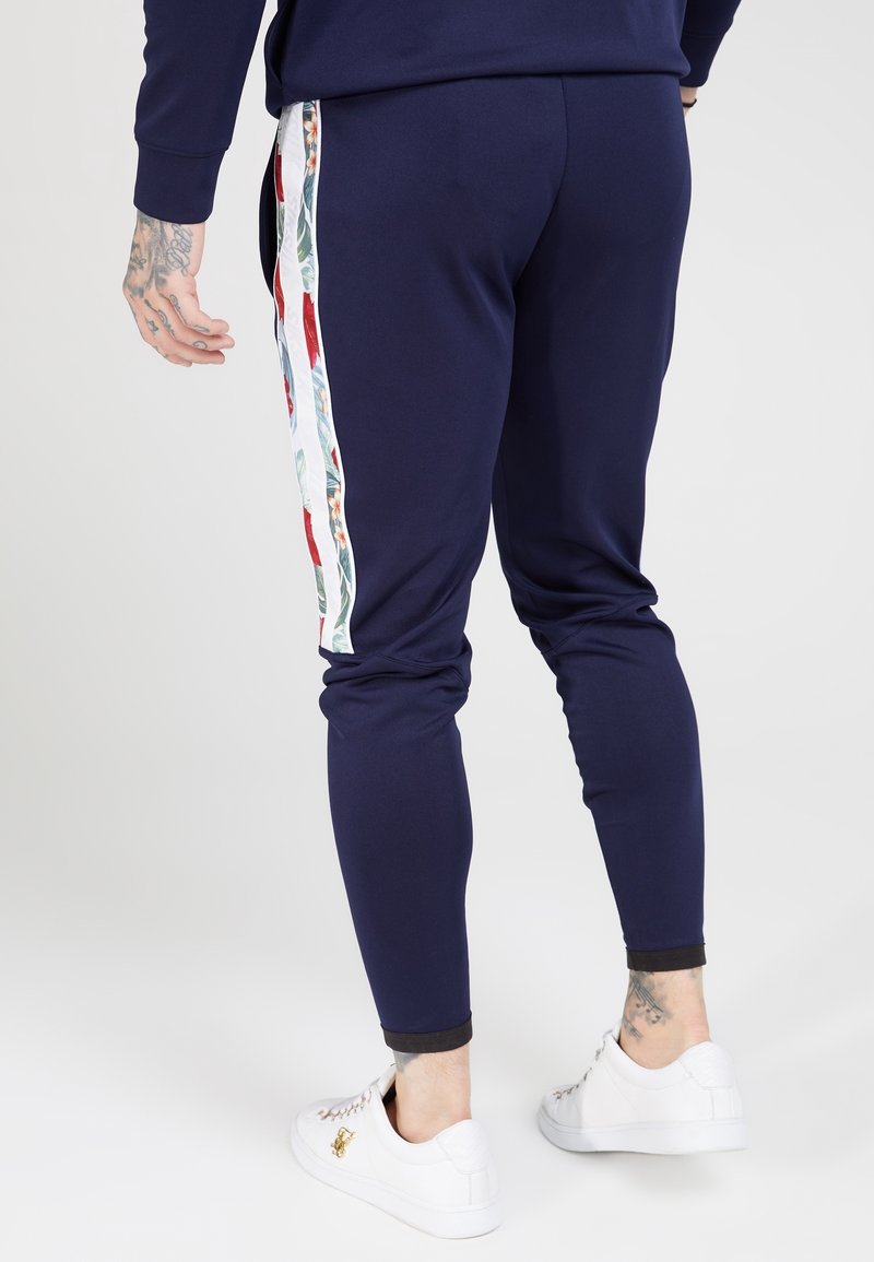 Marineblauwe sportieve joggingbroeken met bloemenrand, aansluitend ontwerp en ribgebreide tailleband. Gepaard met witte sneakers en een zichtbaar tattoo op het been.