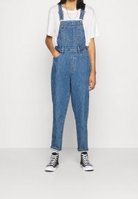 Denimoveraller i mellanblått med justerbara axelremmar, framficka och uppvikta fållar, kombinerat med svartvita högstövlade sneakers.