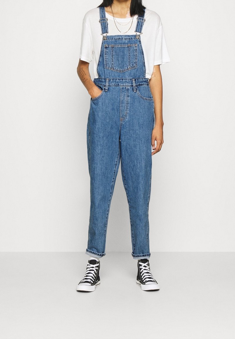 Denimoveraller i mellanblått med justerbara axelremmar, framficka och uppvikta fållar, kombinerat med svartvita högstövlade sneakers.