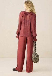 Vrouw met lang golvend blond haar draagt een doorschijnende roestkleurige blouse en wijde broek, terwijl ze een tas met luipaardprint vasthoudt tegen een effen achtergrond.