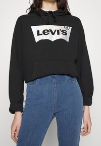 Svart hoodie med en metallisk Levi's-logotyp, utrustad med känguruficka och dragsnodd i huvan, tillsammans med högmidjade blå jeans.