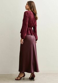 Blusa bordeaux con design floreale testurizzato, vita arricciata e maniche lunghe; abbinata a una gonna liscia in satin bordeaux e scarpe con tacco a striscia.