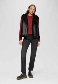 Zwarte faux leren vest met schapenvacht kraag, gecombineerd met een rode kabelgebreide trui, zwarte jeans en chunky zwart-groene enkelboots.