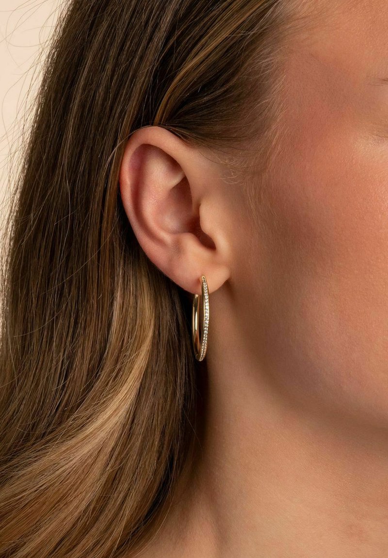 Närbild av en kvinnas öra som visar en guld hoops örhänge prytt med glittrande stenar, med långt, flödande brunt hår som ramar in hennes ansikte.