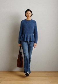 Žena u plavom peplum puloveru, plavim trapericama i bijelim tenisicama, nosi smeđu torbu i hoda po drvenom podu uz običan zid.