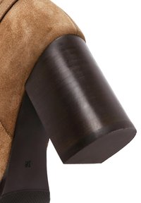 Scarpa con tacco alto in suede marrone, con tacco a blocco in legno scuro e arrotondato e una suola testurizzata. Il tacco presenta delle scanalature sottili per una migliore aderenza.