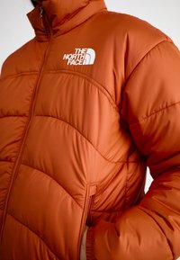 Orange puffer jacket with a zippered front, featuring a stitched pattern and a white logo on the left side. Includes side pockets.