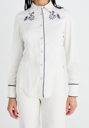 Hvid langærmet skjorte med navyblåt blomsterbroderi på skuldrene, navy piping langs knaprækken og manchetterne, båret med hvide bukser.