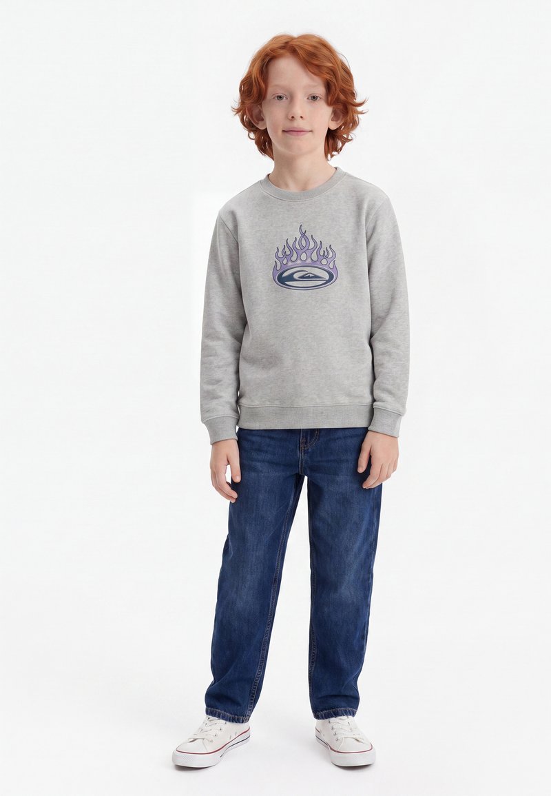 Garçon aux cheveux roux portant un sweat-shirt gris avec un motif de flammes, un jean bleu et des baskets blanches, debout sur un fond blanc.