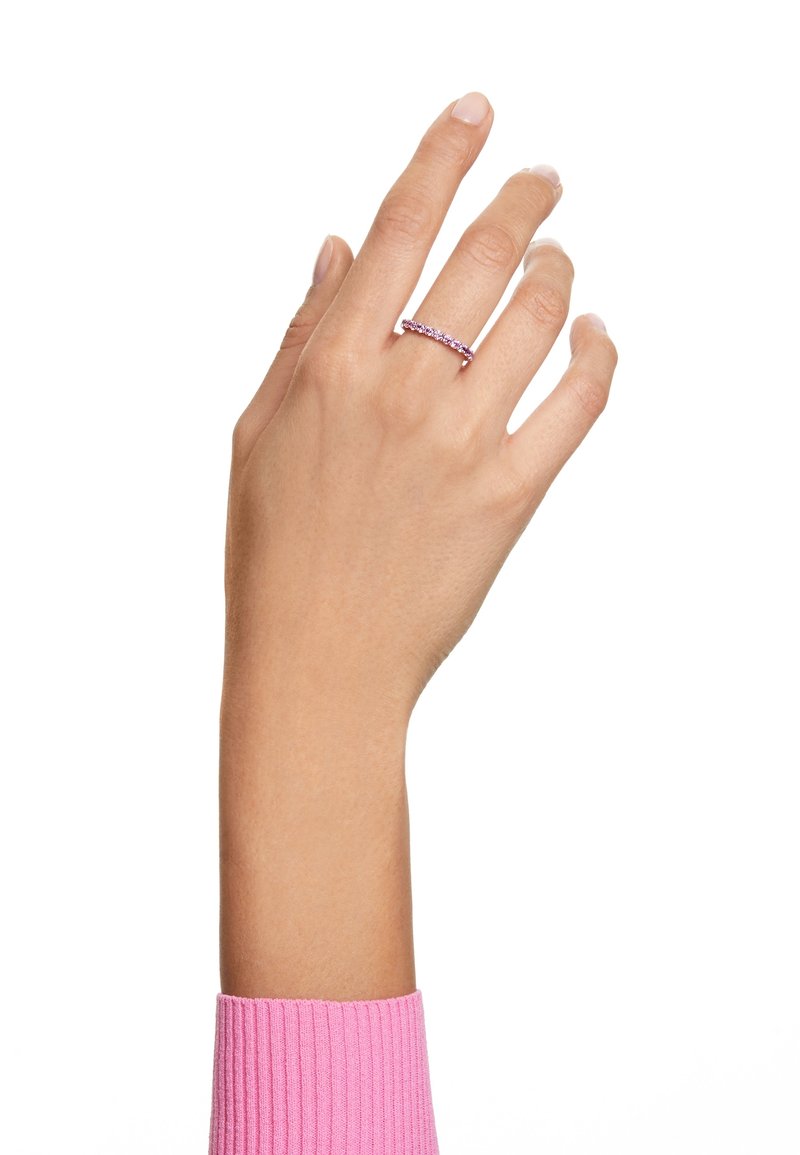 Main portant une bague à pierre précieuse rose au majeur, levée sur un fond blanc, avec un poignet côtelé rose visible.