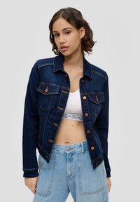 Denimjacket in donkerblauw, met twee borstzakken, koperen knopen en contrasterende stiksels. Model draagt eronder een witte tanktop.