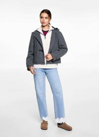 Grijze cropped jas met kap, voorzien van een dubbelzijdige sluiting, gedragen over een lichte trui met rits, gecombineerd met lichtblauwe jeans en bruine schoenen.