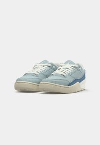 Baskets en denim bleu avec des lacets blancs, présentant une tige texturée et des accents d'un bleu plus sombre, ainsi qu'une semelle en caoutchouc beige.