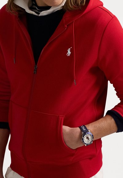 Fle hoodie vermelho com fecho éclair, feito de um tecido suave, com um bolso frontal e um logótipo bordado. Usado por cima de um suéter azul-marinho com um relógio no pulso.