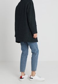 Abrigo texturizado negro, con un corte ligeramente oversize y mangas largas. Pantalones vaqueros ajustados de color azul claro y zapatillas blancas con acentos rojos.