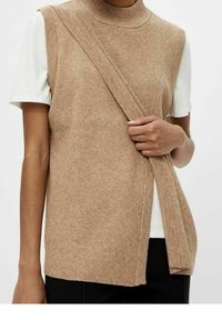 Femme portant un gilet en tricot beige sans manches sur une chemise blanche à manches courtes, tenant le bord avant du gilet d'une main.